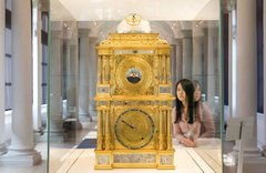 Besucherin betrachtet große, goldene, reich verzierte Uhr mit zwei Ziffernblättern auf Vorderseite